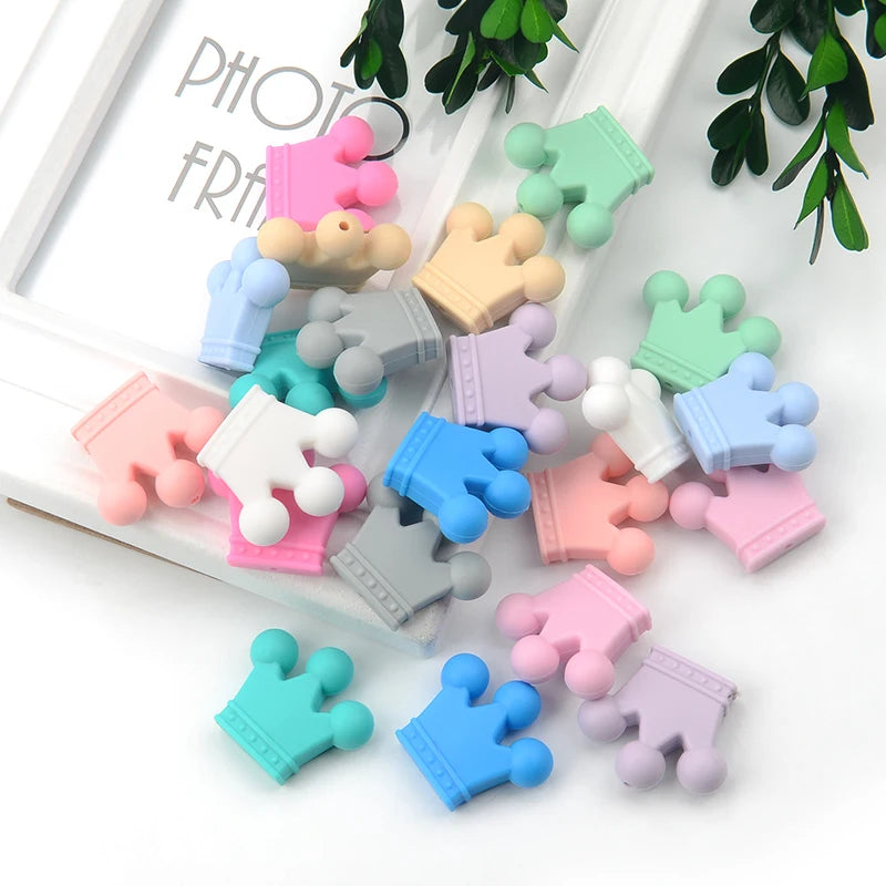Colorful crown-shaped silicone beads scattered on a white surface, surrounded by green leaves and partially covering a white photo frame with some text visible.