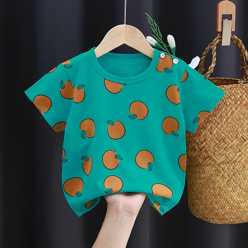 Bright teal children's t-shirt with orange fruit pattern, featuring button details on the shoulder. It hangs on a wooden hanger beside a woven basket and dark background.