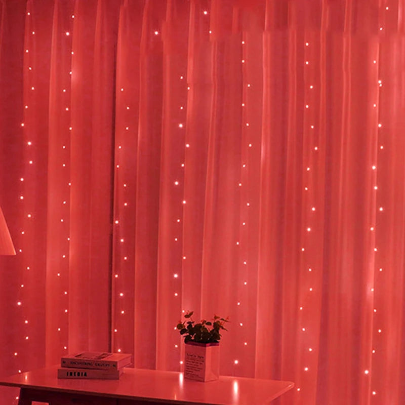 A softly lit room with red curtains decorated by twinkling fairy lights. A table with books and a small potted plant sits in front, next to a floor lamp.