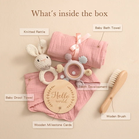 A collection of baby items includes a pink bath towel, drool towel, knitted rattle, teething toys, a wooden hairbrush, and milestone cards with "Hello world" text, all elegantly arranged.