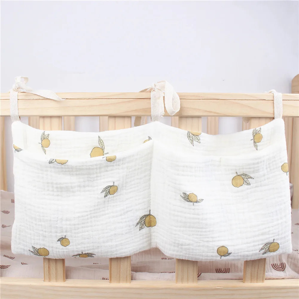 White cloth basket with lemon prints attached to a wooden crib, using tied fabric loops. The basket has two compartments and is designed for storing baby's items.