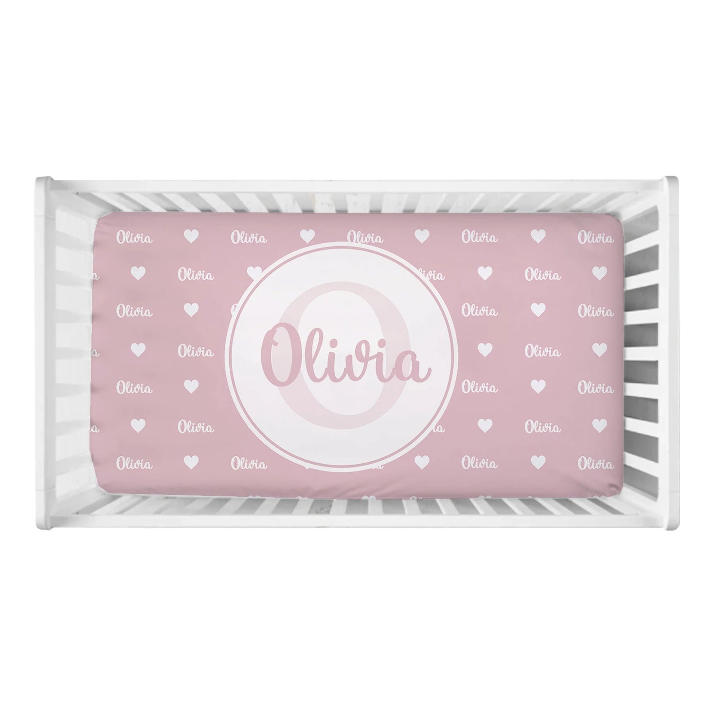 White crib with a pink mattress featuring "Olivia" in white, surrounded by small white hearts. A large circle in the center highlights the name in script font.