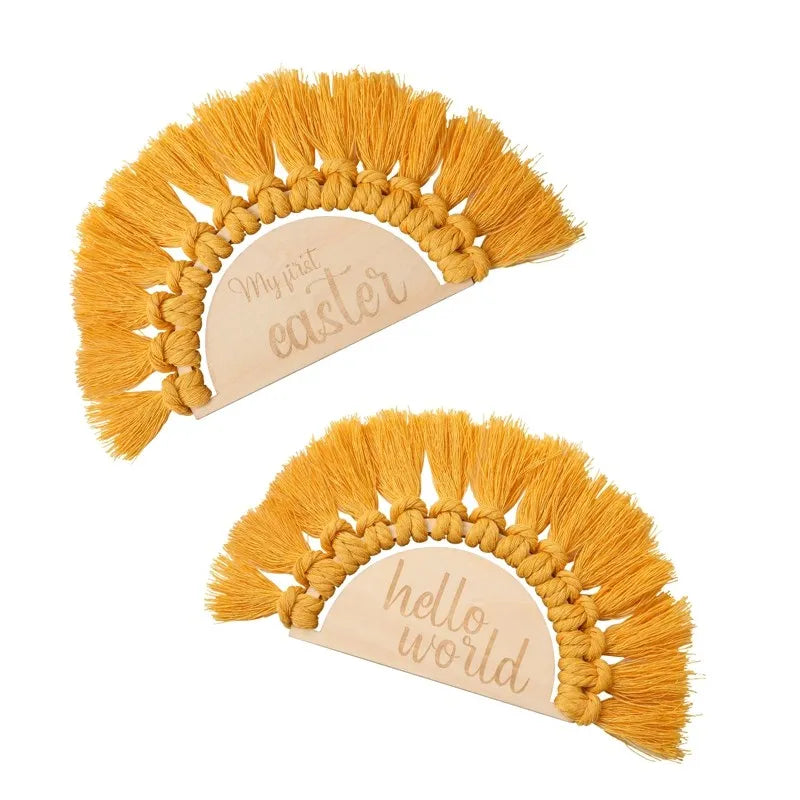 Two semi-circular wooden plaques with mustard macrame tassels. One reads "My first easter," and the other says "hello world." Both are decorative and cheerful.