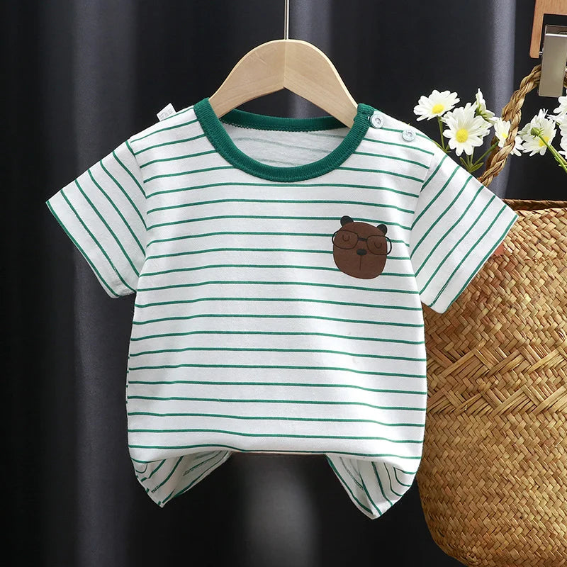 Striped baby t-shirt with green trim, featuring a cute bear face graphic. Hangs on a wooden hanger against a dark curtain, next to a woven basket with white flowers.