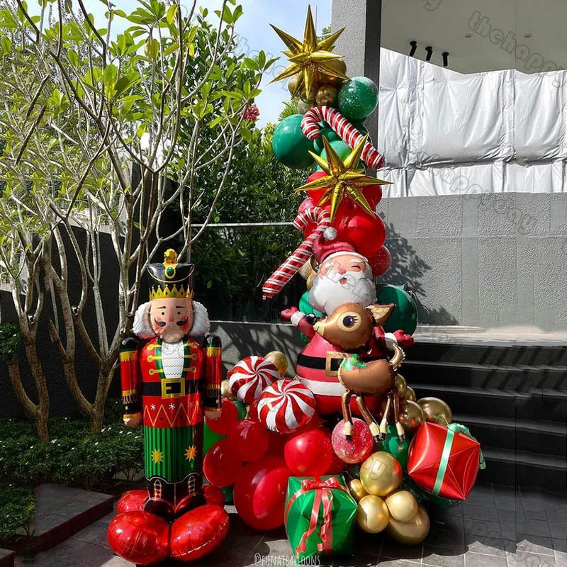 Festive outdoor scene with a nutcracker, Santa, reindeer, candy cane, stars, and gift balloon sculptures. Decorated in red, green, and gold near a concrete staircase and leafy tree.