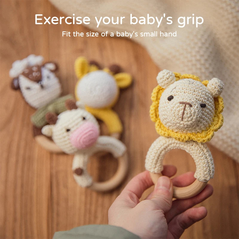 Hand holding a crocheted lion baby rattle with a wooden ring. Three other animal rattles lie on a wooden surface. Text above: "Exercise your baby's grip. Fit the size of a baby's small hand."