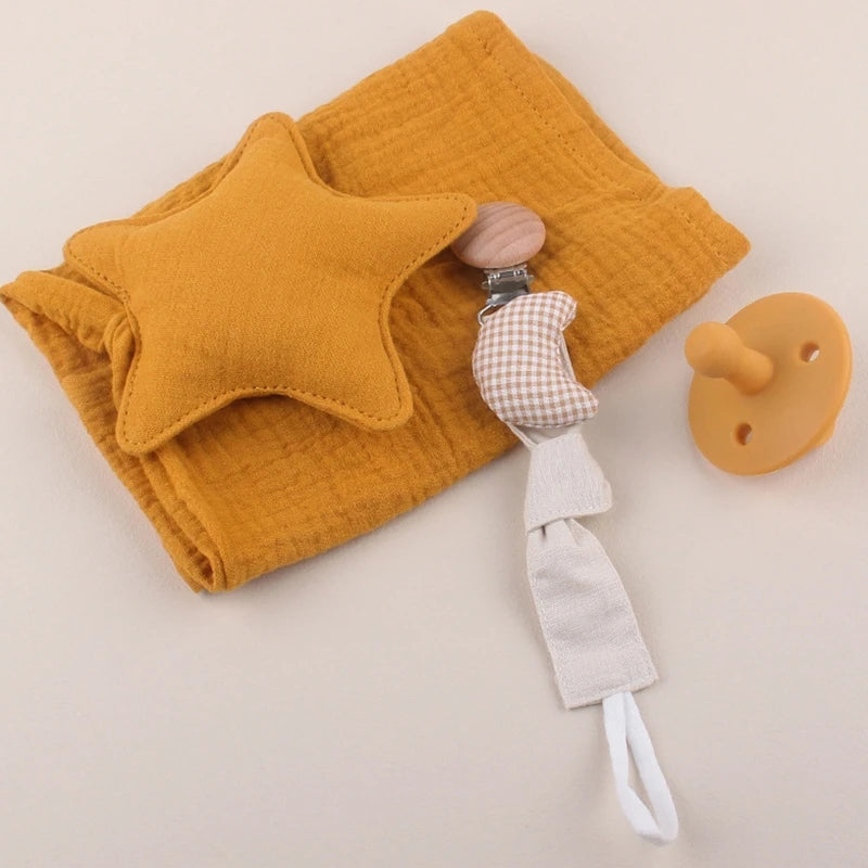 A mustard muslin cloth with a star cushion, a gingham pacifier clip, and a mustard pacifier are arranged on a light surface.