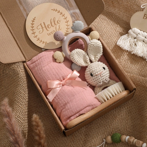 Gift box with a pink muslin blanket, plush bunny toy, wooden brush, teething ring, and "Hello World" sign. Set against a burlap background with natural decor accents.