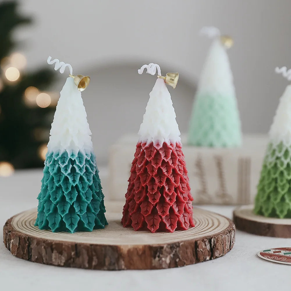 Two textured, cone-shaped candles resembling trees, one in teal and white, the other in red and white, are topped with small gold bells and displayed on wooden slabs.