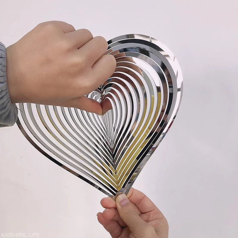 Two hands holding a metallic, heart-shaped wind spinner with layered, reflective strips creating an optical illusion against a plain background.