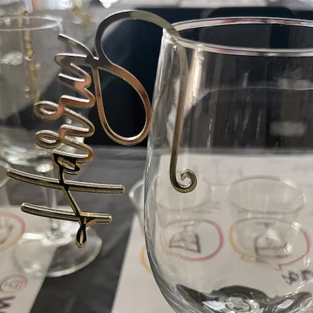 A metal name tag reading "Harry" elegantly hangs on the rim of a clear wine glass, enhancing a sophisticated table setting with its cursive design and reflective surface.