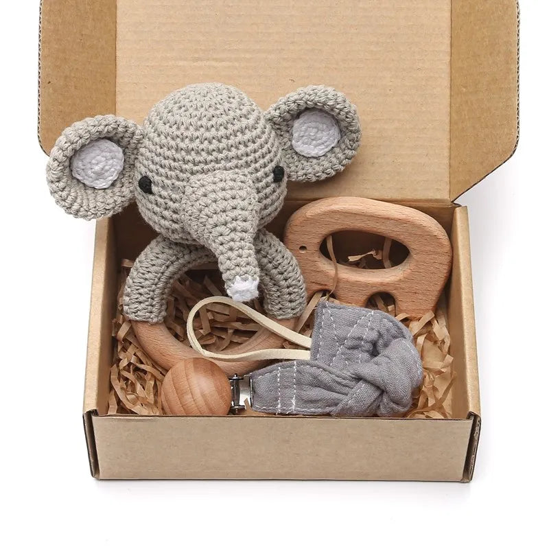 A gift box containing a crocheted elephant toy, a wooden teething toy, and a fabric pacifier clip, all nestled in shredded brown paper.