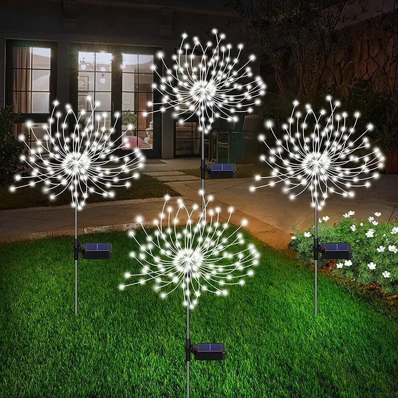 Solar-powered dandelion-shaped LED lights illuminate a garden pathway at night, casting a warm glow on the grass and nearby flowers in front of a house.