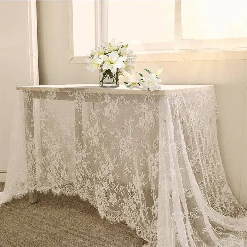 A white table draped with floral lace fabric stands in front of a window. A vase of white lilies sits on top, adding a touch of elegance.