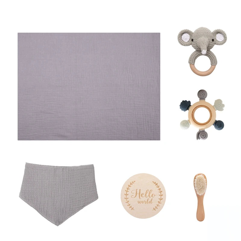 Baby gift set includes a gray muslin blanket, knitted elephant rattle, wooden teether, teething ring, gray bib, engraved "Hello World" wooden disc, and a soft baby brush.