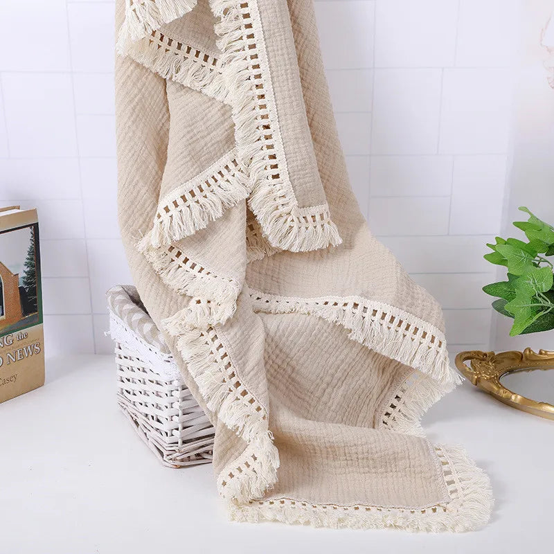 Cream-colored textured blanket with fringe edges draped over a wicker basket on a white surface. A green plant and a gold-framed mirror are nearby.