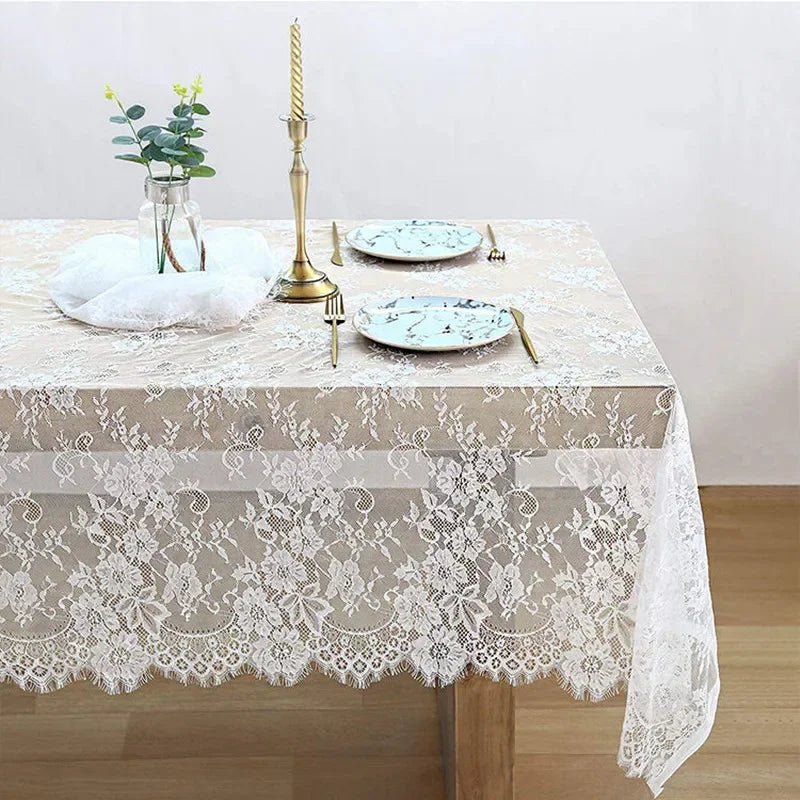 Elegant table setting with a floral lace tablecloth, gold cutlery, marble-patterned plates, a tall brass candle holder, and a glass vase with greenery on a wooden floor.