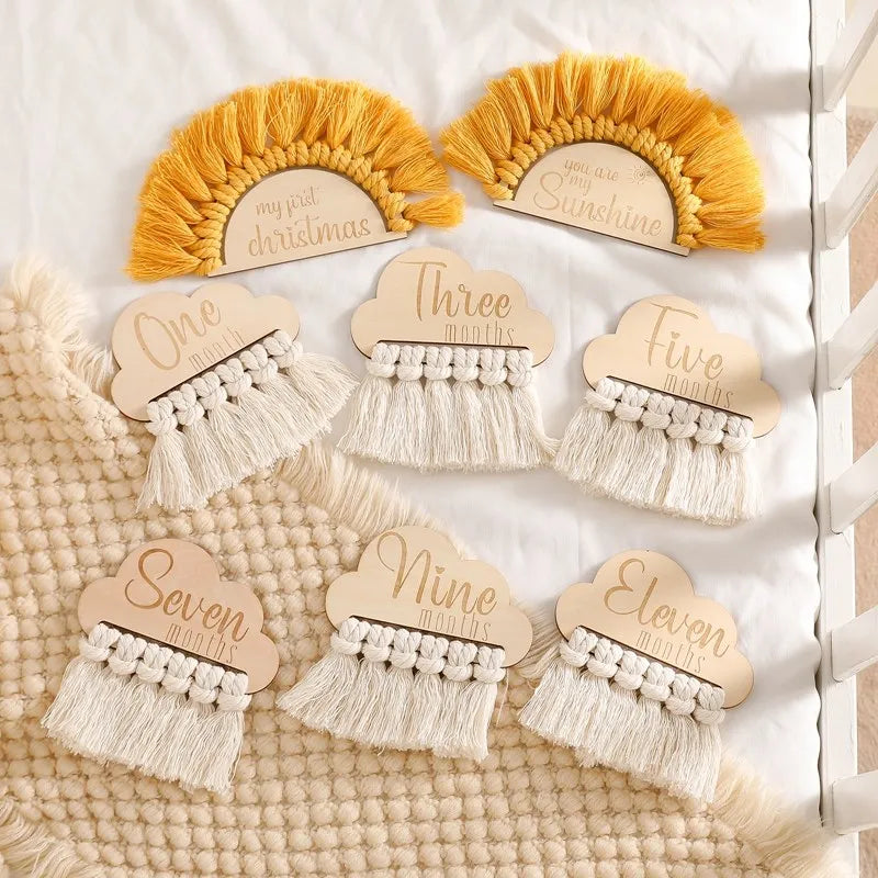 Wooden milestone plaques with months and events, decorated with woven tassels, on a textured blanket. They celebrate baby's growth and special moments.