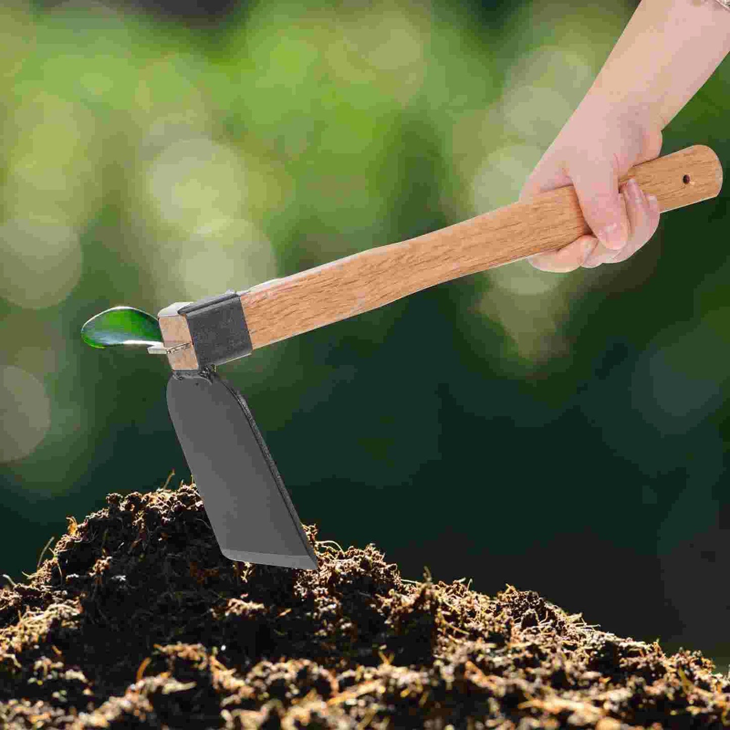 A hand uses a wooden-handled hoe to till rich, dark soil. A small green sprout is visible, surrounded by a blurred green background, indicating a garden setting.
