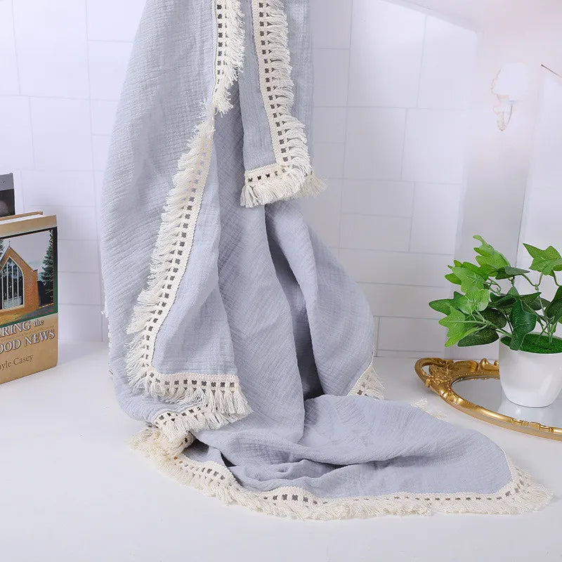 A soft, light gray blanket with decorative lace edges draped on a white surface. A book and a small green potted plant are nearby, adding a cozy and elegant touch to the scene.