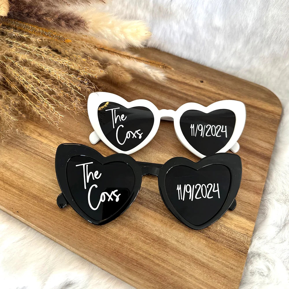 Heart-shaped sunglasses in black and white featuring text "The Coxs" and date "11/9/2024" on the lenses, resting on a wooden surface with dried pampas grass nearby.