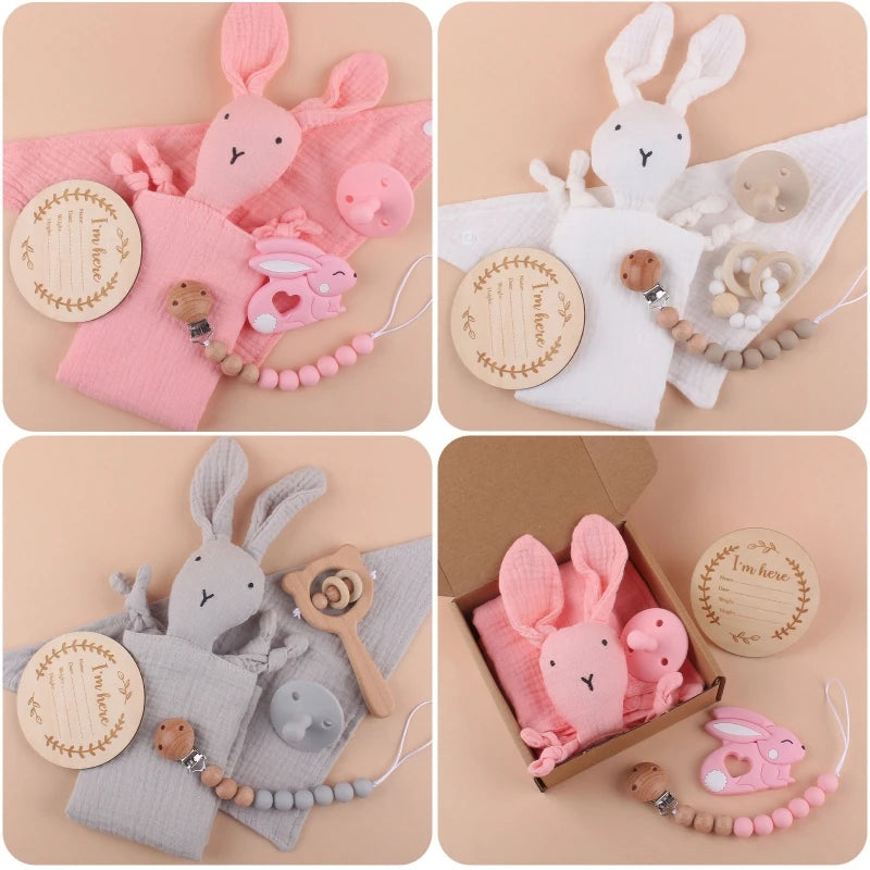 Baby gift sets featuring soft bunny toys in pink, white, and gray. Includes wooden plaques, teething rings, clips, and pacifiers. One set shown in a brown box.