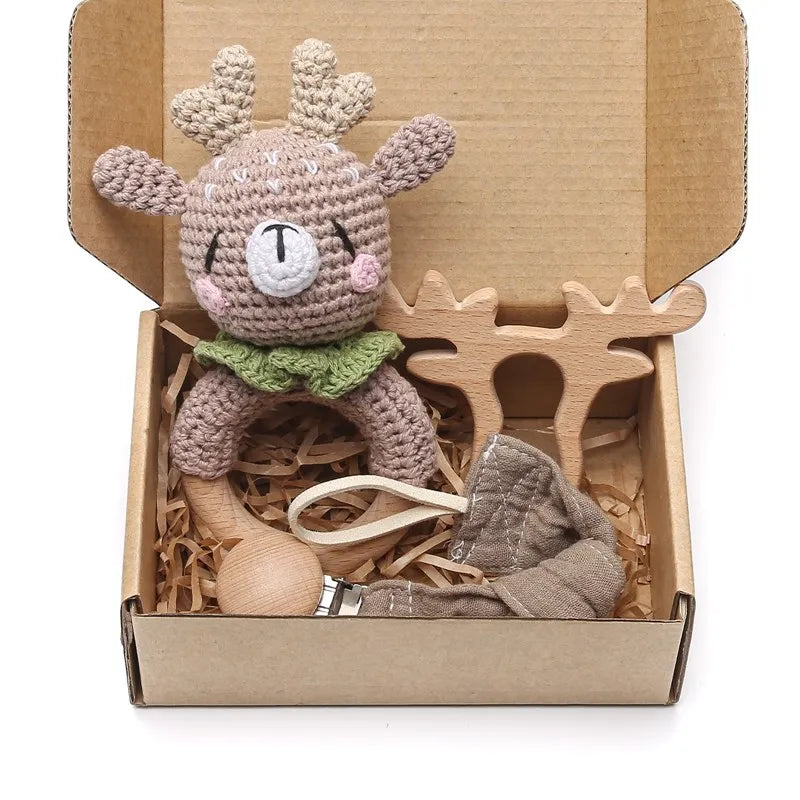 A gift box containing a crocheted deer toy with antlers and a green collar, a wooden reindeer-shaped teether, and a fabric pacifier clip on shredded paper bedding.