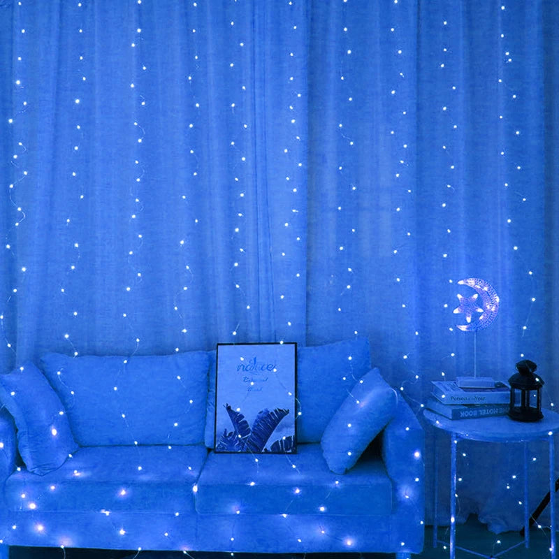 Blue room with fairy lights on curtains, a blue sofa with cushions and a framed picture. A side table holds books, a lamp with moon design, and a small lantern.