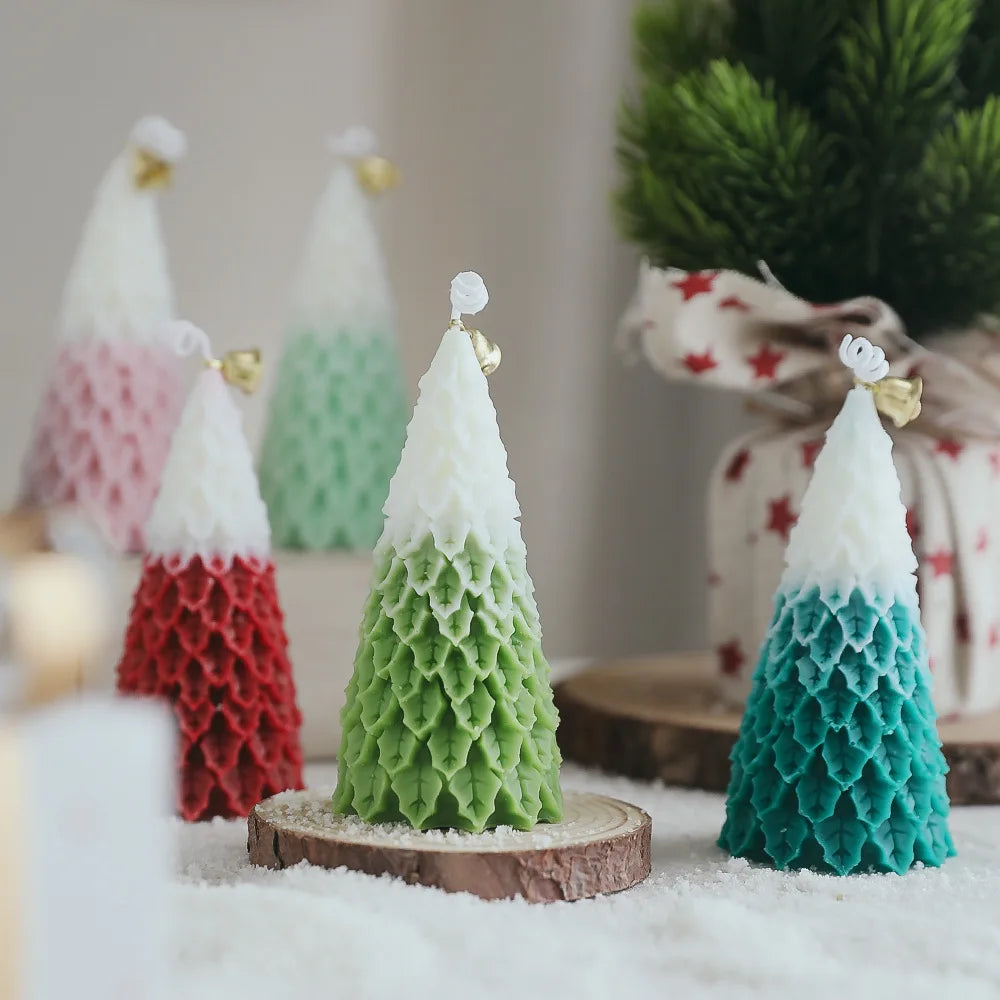 Festive, tree-shaped candles with textured layers in shades of green, red, pink, and teal. Each candle has a white top, adorned with a small gold bell, set on a snowy surface.