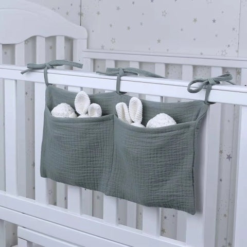 Soft green fabric organizer with two pockets hangs on a white crib, holding baby essentials like plush toys and rolled cloths. Background features a wall with small star patterns.