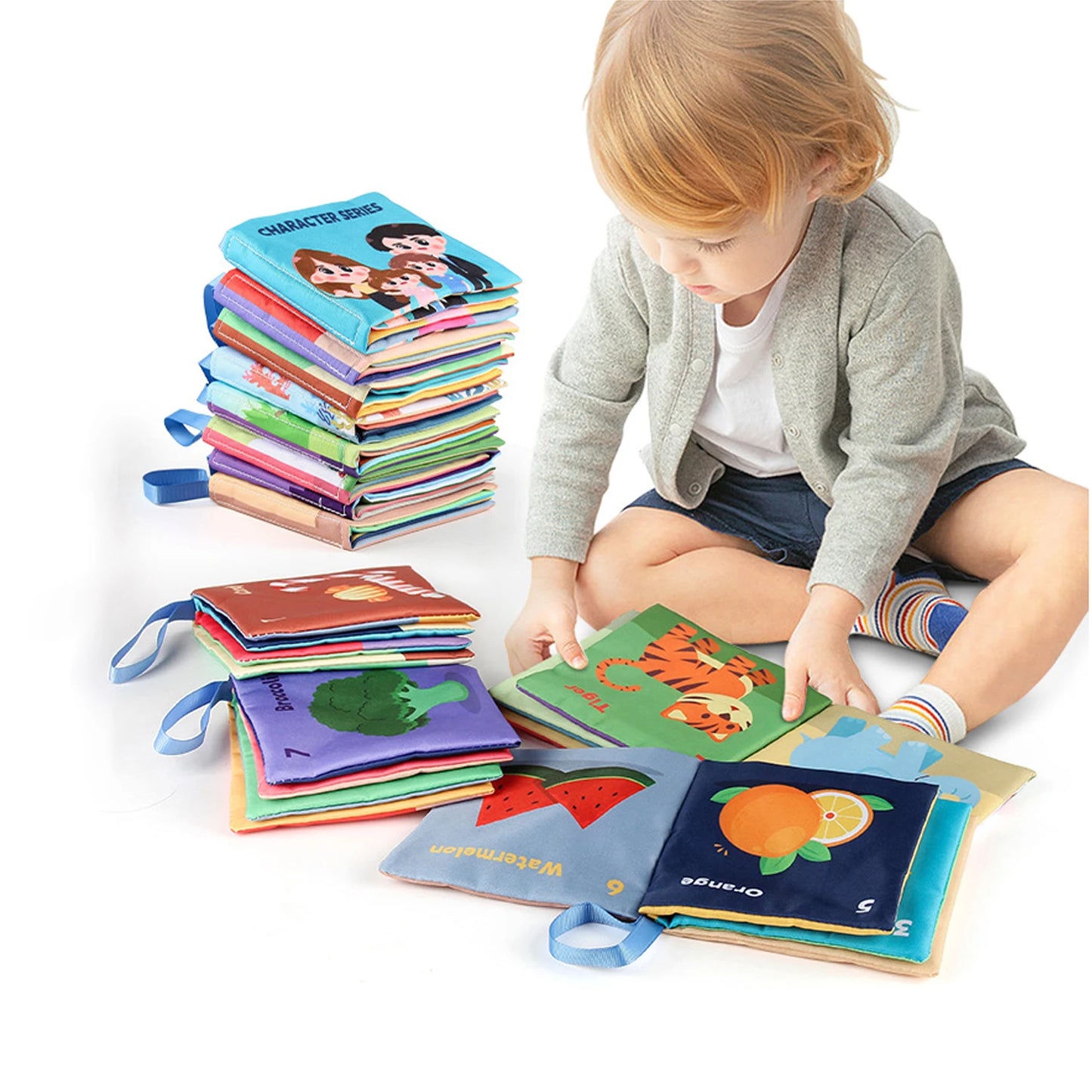 A young child sits cross-legged on the floor, exploring colorful fabric books with illustrations of animals and fruits. A tall stack of similar books is nearby.