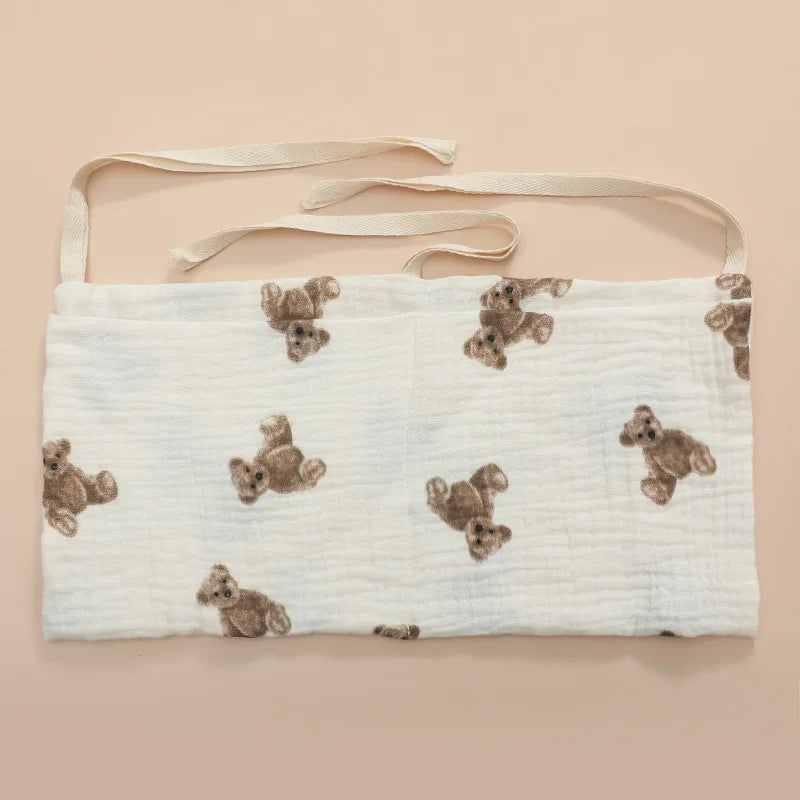 White fabric apron with cute brown teddy bear print, featuring two long ties at the top for fastening. The background is a soft beige color.