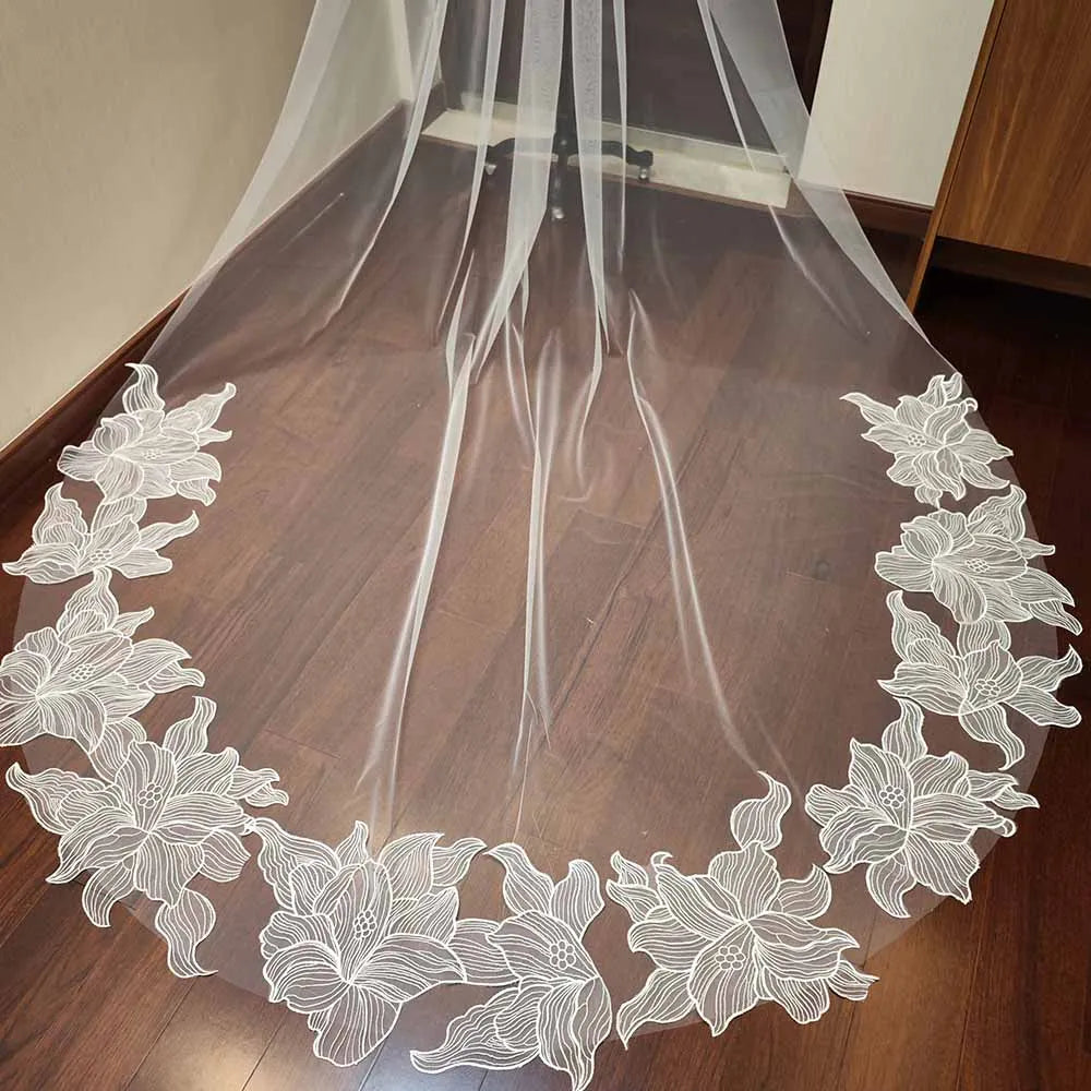 Sheer white curtain with an embroidered floral border in delicate patterns, drapes over a polished wooden floor, creating a soft and elegant look.