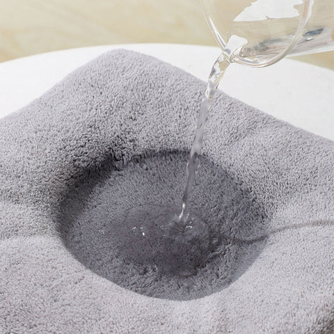 Water is being poured from a glass onto a soft, gray, absorbent mat, creating a wet, darkened area in the center. The surface beneath is light-colored.