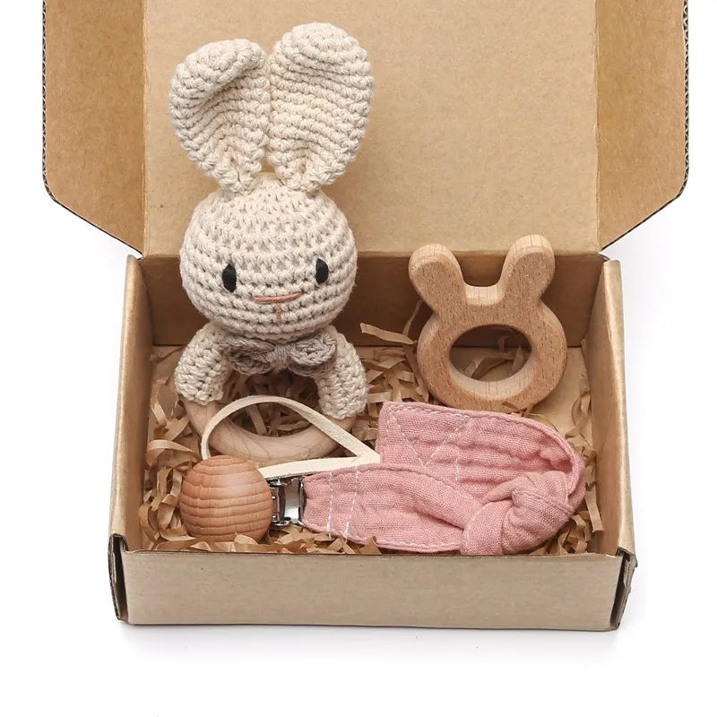 Crochet bunny toy, wooden teether, pacifier clip, and pink cloth in a brown gift box filled with shredded paper.