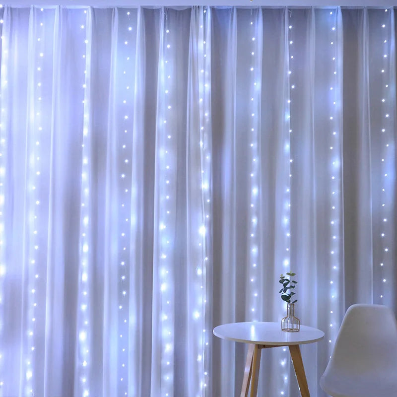 White curtain with cascading fairy lights creating a dreamy glow. A small round table with a vase of green leaves and a white chair are in the foreground.