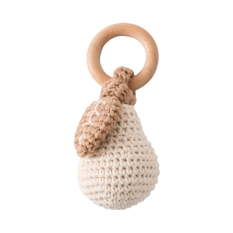 Crocheted pear-shaped baby rattle with a wooden ring handle and knitted leaf detail. The rattle is soft beige with a light brown leaf, combining yarn and wood materials.