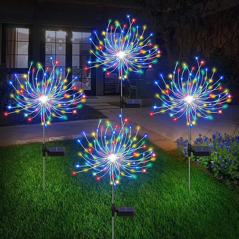 Four colorful LED garden lights resembling fireworks are illuminated on a yard at night. Each light has multicolored bulbs arranged in a circular pattern, creating a festive atmosphere.