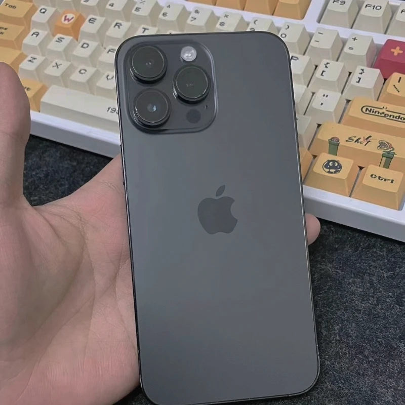 A hand holding a black smartphone with three camera lenses, logo visible. In the background, a colorful mechanical keyboard featuring custom keycaps.