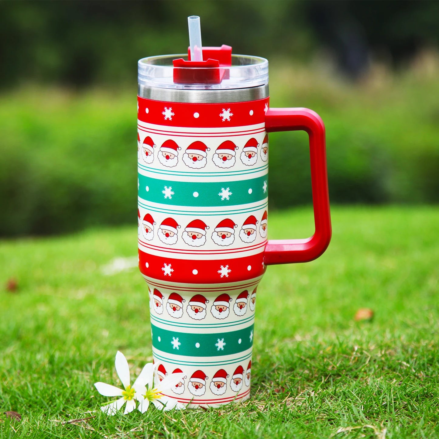 Festive tumbler with Santa designs, red and green stripes, a red handle, and straw lid, sits on grass beside two white flowers, with blurred greenery in the background.