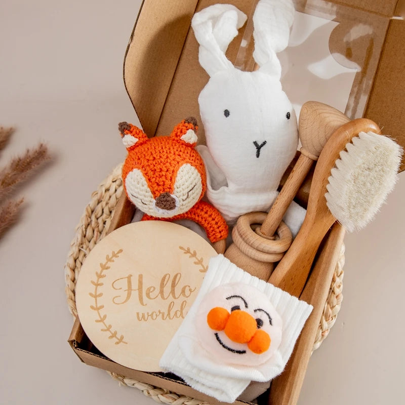 Gift box with a crocheted orange fox, white bunny plush, wooden hairbrush, baby socks with face design, and a round wooden plaque with "Hello world" engraving.