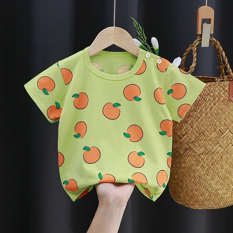 A lime green baby T-shirt with an orange fruit pattern, featuring small green leaves. It's displayed on a wooden hanger with a woven basket in the background.