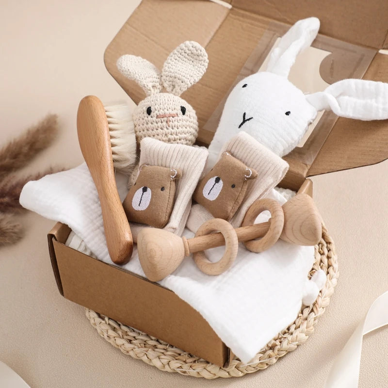 A gift box contains a soft woven knit bunny, white fabric bunny, wooden hairbrush, rattles, and baby socks with bear faces. It is placed on a woven mat with a soft blanket.
