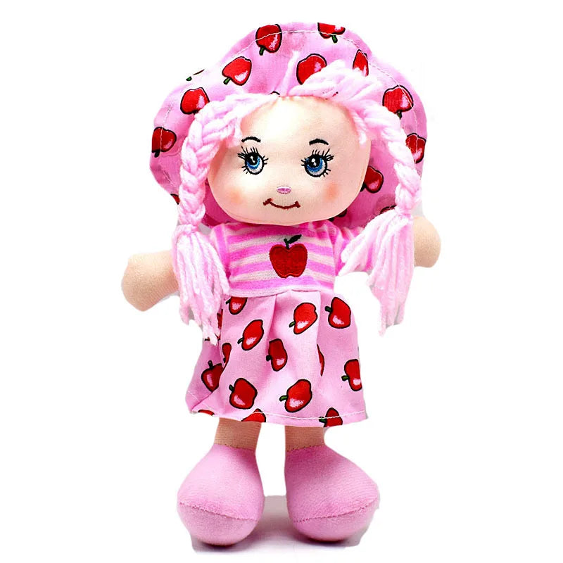 A soft doll with pink braided hair, wearing a pink dress and hat with a red apple pattern. The doll has embroidered facial features and pink shoes.