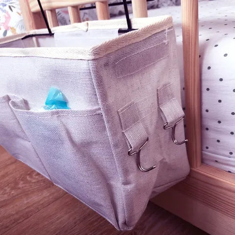 Hanging fabric organizer attached to a wooden bed frame, featuring a small pocket with a blue item. The organizer has hooks and straps for secure attachment.