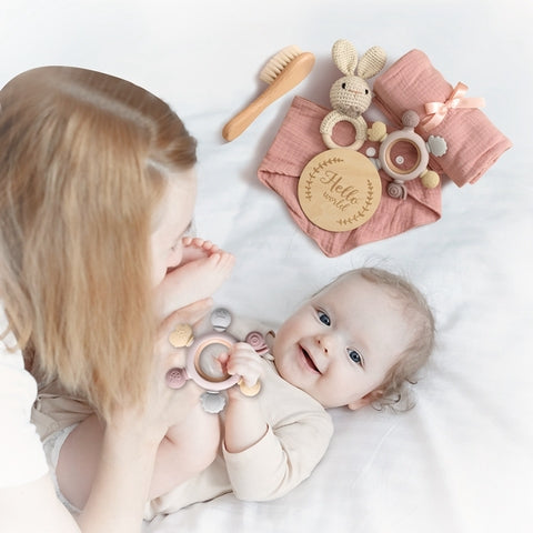 A mother cuddles her smiling baby, who holds a soft rattle. Beside them are baby essentials: a brush, pink blanket, crocheted bunny, and a wooden sign saying "Hello."