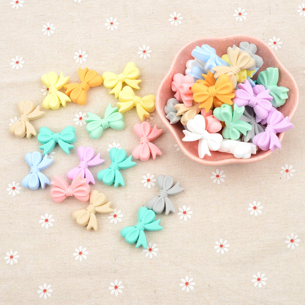 A collection of colorful, small bow-shaped embellishments in pastel shades scattered and in a pink ceramic bowl, on a light beige fabric with white floral patterns.