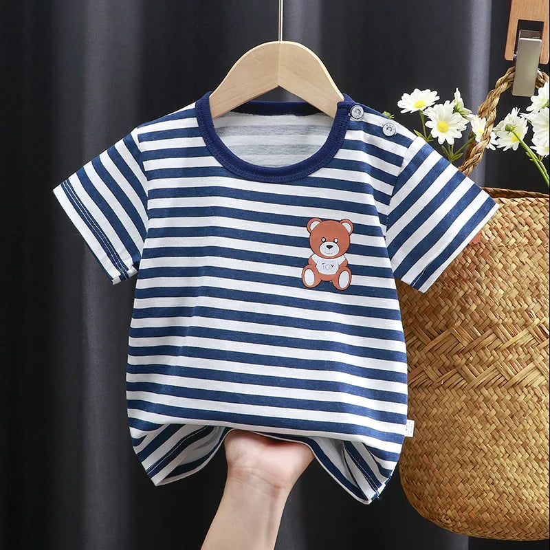 Striped blue and white baby shirt with a teddy bear patch, held by a hand. Shirt is on a hanger next to daisies and a woven basket, with a dark background.