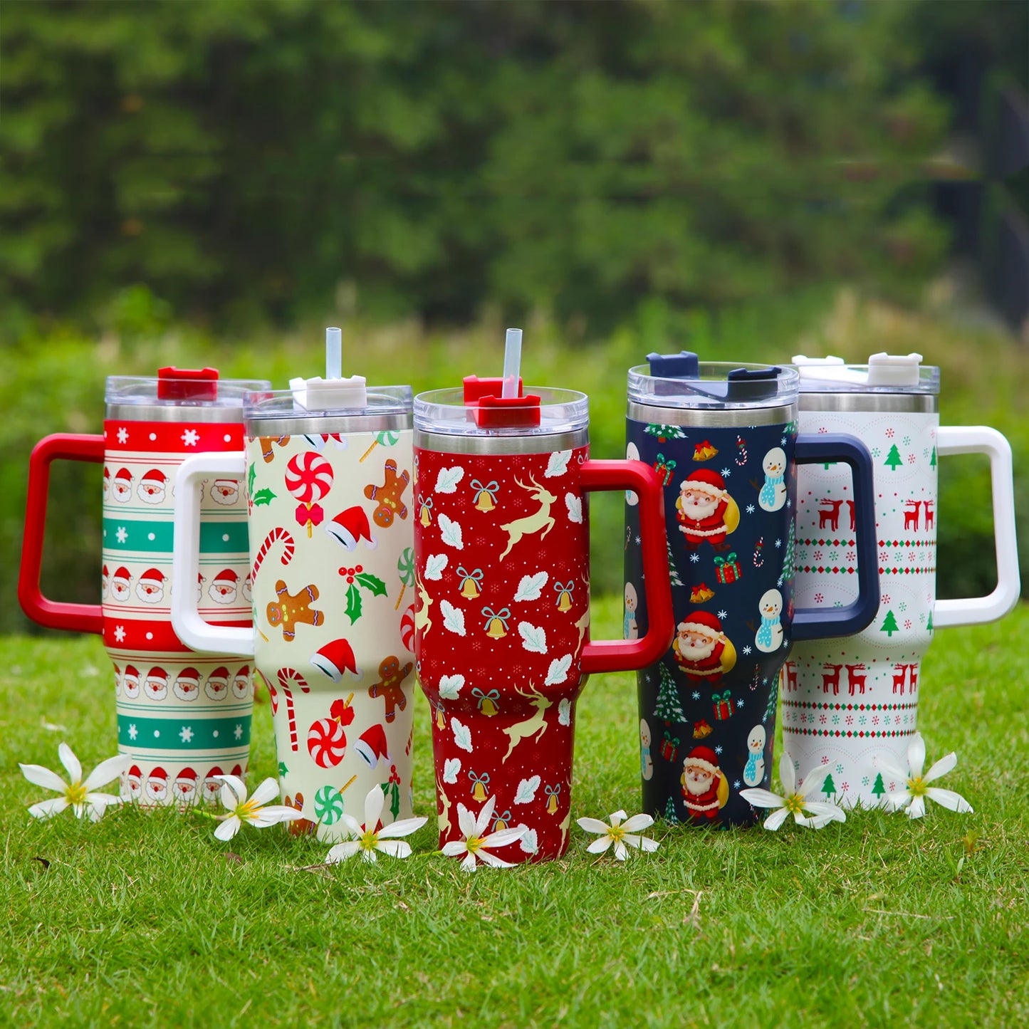 Five festive tumblers with Christmas designs, including Santa, gingerbread, and reindeer, sit on grass with small white flowers. Each has a large handle and straw lid.