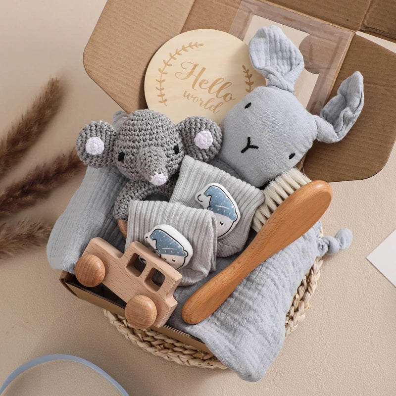 Gift box with gray stuffed bunny, knitted mouse, baby socks, wooden toy car, hairbrush, and a round "Hello World" card on a beige background.